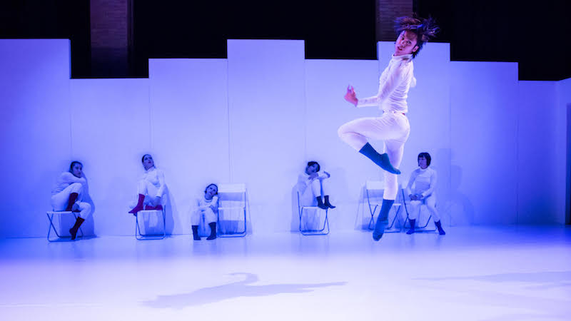 Dancers dressed in white save for colorful socks sit against a wall while another jumps into the air looking over their shoulder